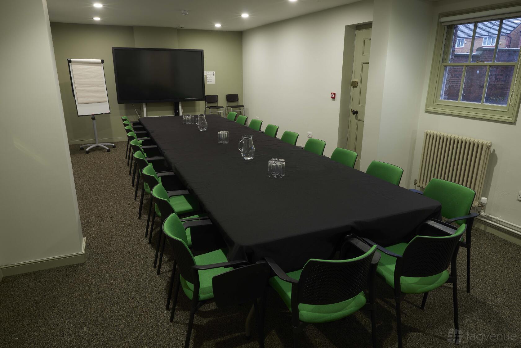 A meeting room with a long table covered in a black cloth, green chairs, a flip chart, and a large screen at Elizabeth Gaskell's House.