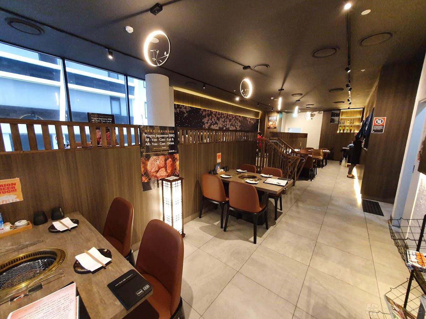 A restaurant with wooden tables, brown chairs, built-in grill tops, and warm ambient lighting at Kuro Sakura Japanese BBQ.