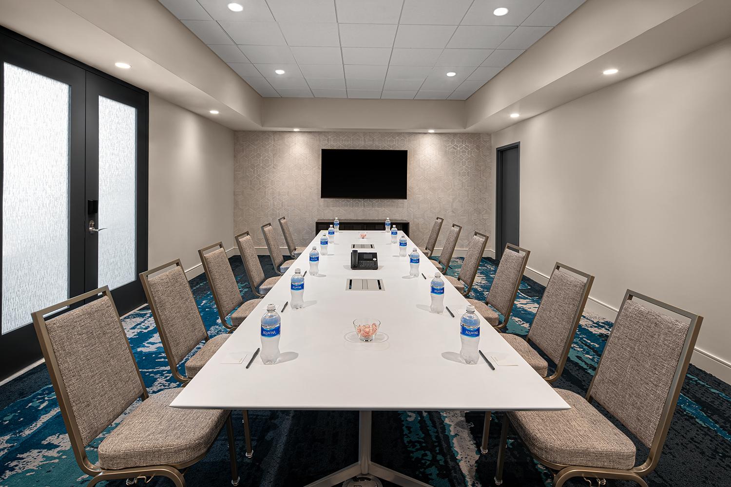 A boardroom with a long rectangular table, bottled water, notepads, and a wall-mounted TV at Courtyard Delray Beach.