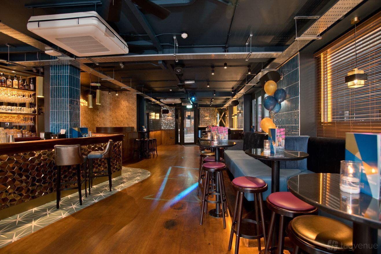 A cocktail bar with high tables, blue-tiled columns, wooden floors, and a stocked bar at Be At One Farringdon.