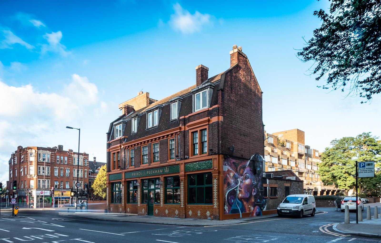 A corner pub with a brick facade, large street-facing windows, and a mural on the side at Prince of Peckham.