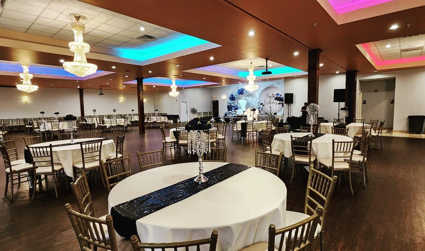 A banquet hall with round tables covered in white linens, gold chairs, and colorful ceiling lighting at Minerva Banquet Hall.