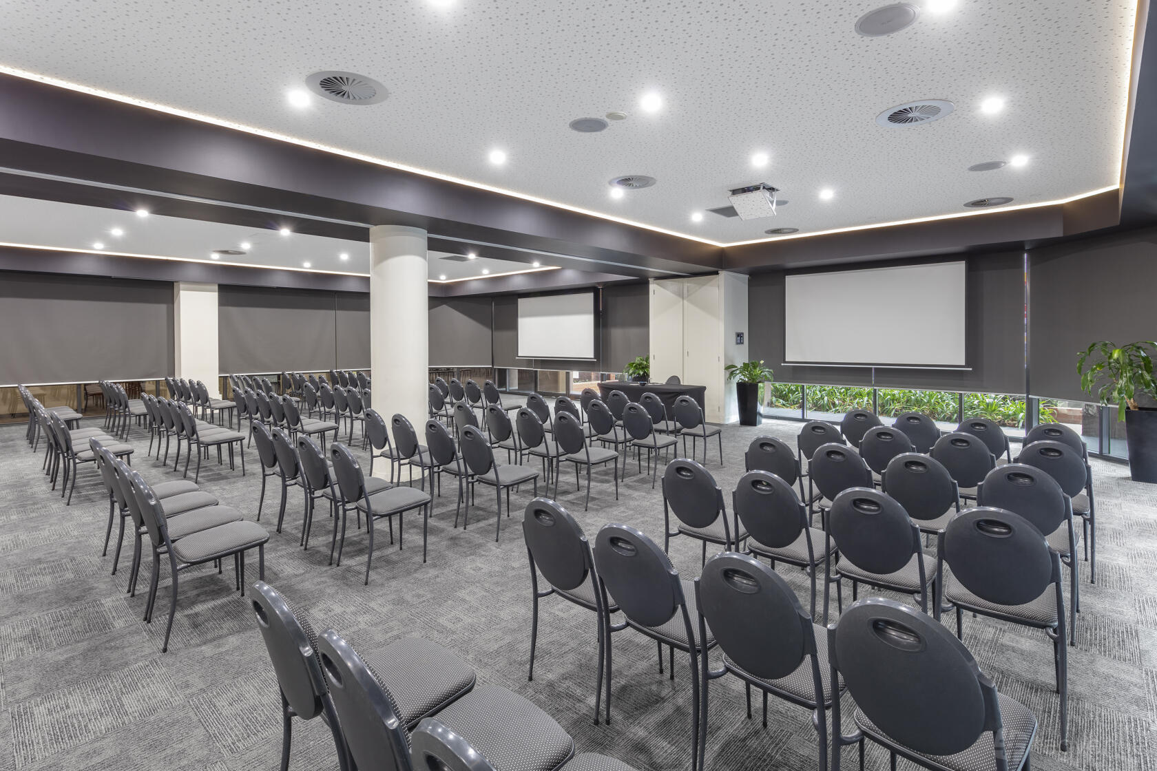 An event space with rows of black chairs, ceiling-mounted projectors, and large windows at Amora Hotel Riverwalk Melbourne.