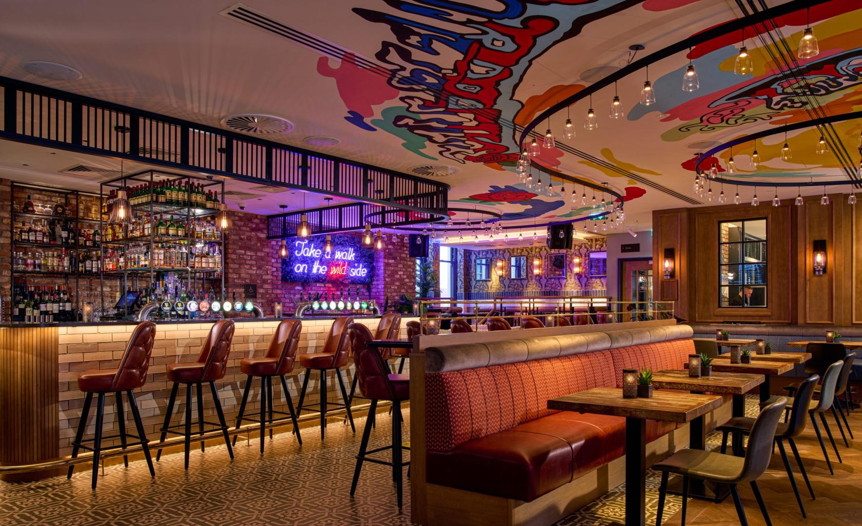 An event space in a hotel with colorful ceiling murals, a neon sign above the bar, and red upholstered booths at NYX Bar & Restaurant.