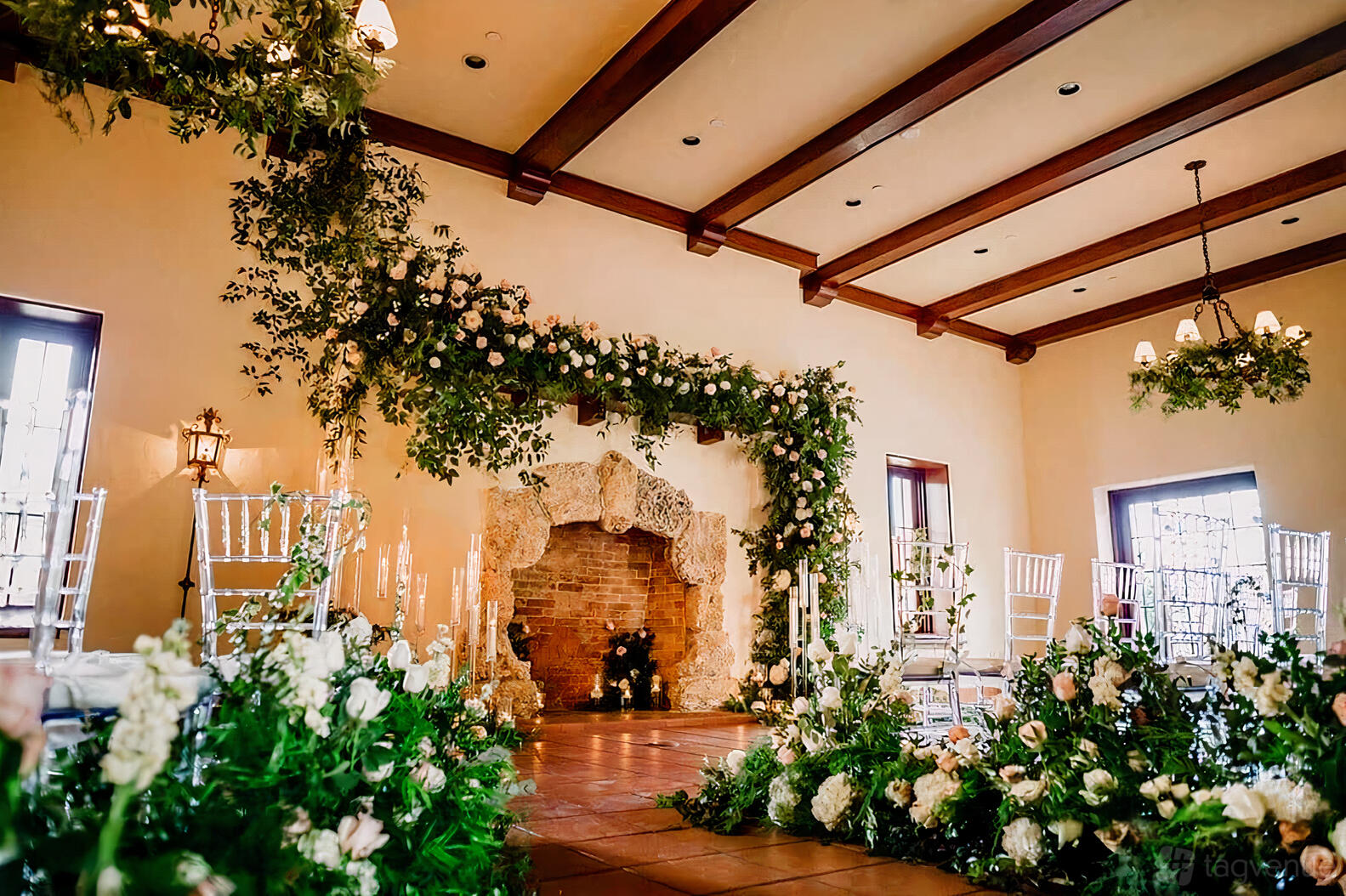 An event venue with exposed wood ceiling beams, a stone fireplace, and floral arrangements at Curtiss Mansion.