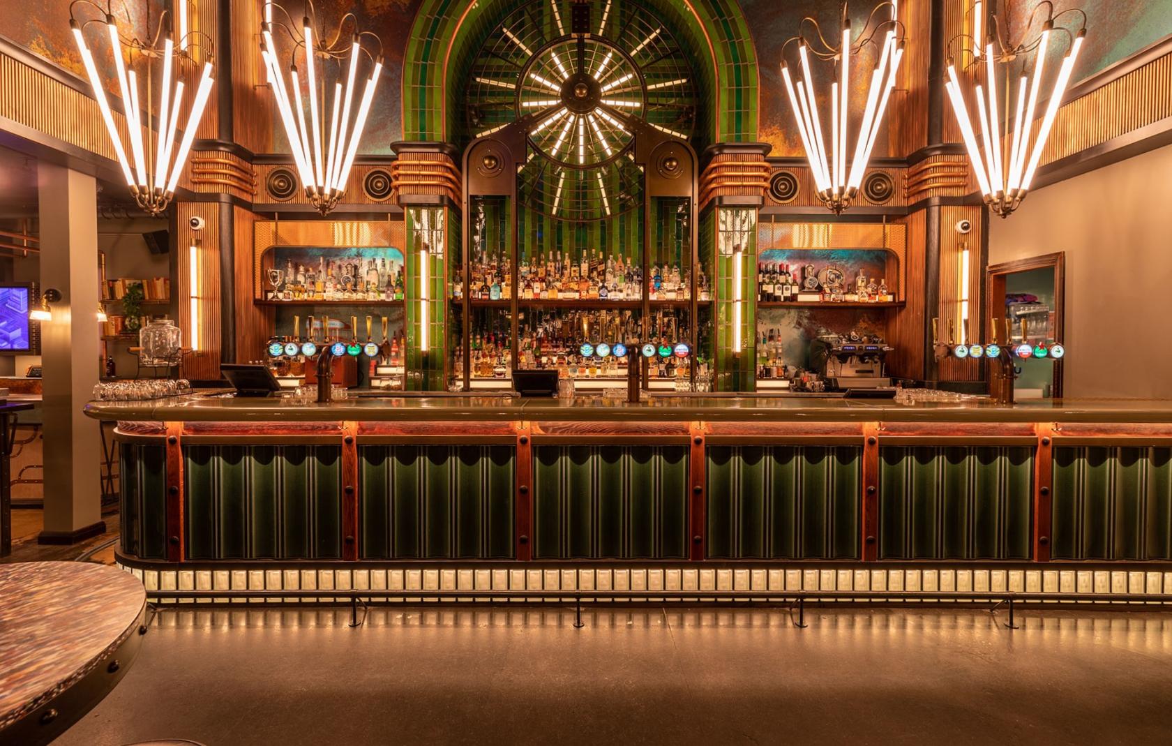 A bar with an illuminated counter, art deco lighting fixtures, and a mirrored back bar at Electric Shuffle Canary Wharf.
