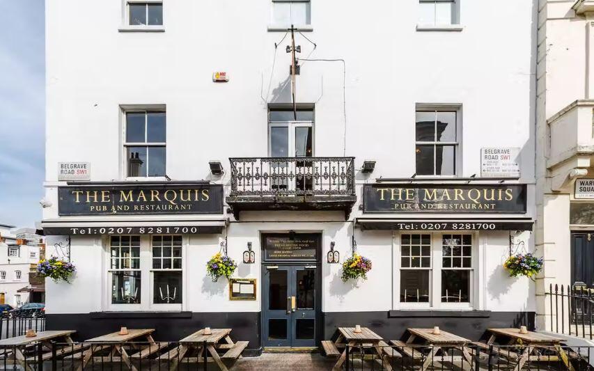 A classic pub with a white facade, hanging flower baskets, black signage, and outdoor wooden benches at Marquis of Westminster.