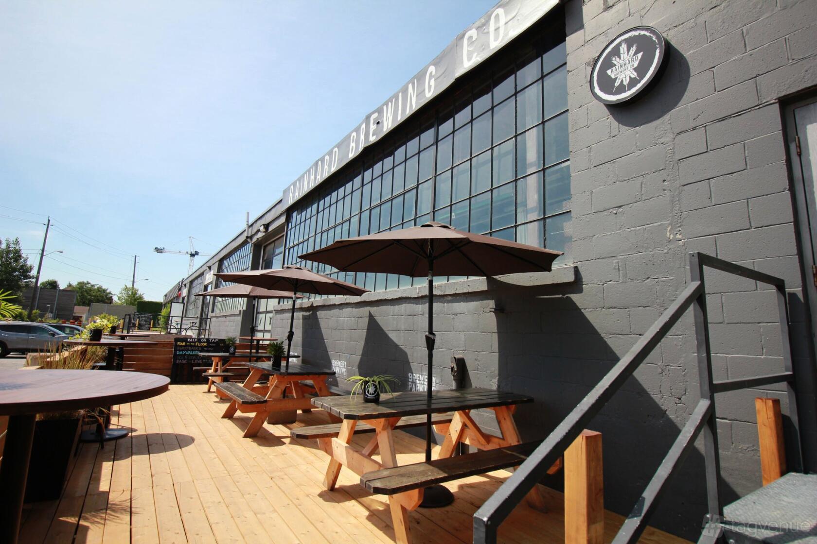 An outdoor patio with wooden picnic tables and umbrellas along a brewery's industrial-style exterior at Rainhard Brewing Co.