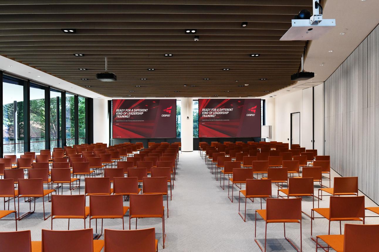An auditorium with rows of orange chairs, floor-to-ceiling windows, and dual projection screens at Catapult.