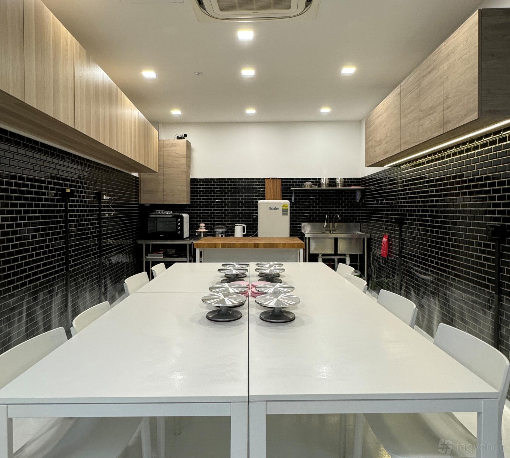 A kitchen with white tables, cake turntables, black tile backsplash, and stainless steel appliances at Meatballs baking studio.