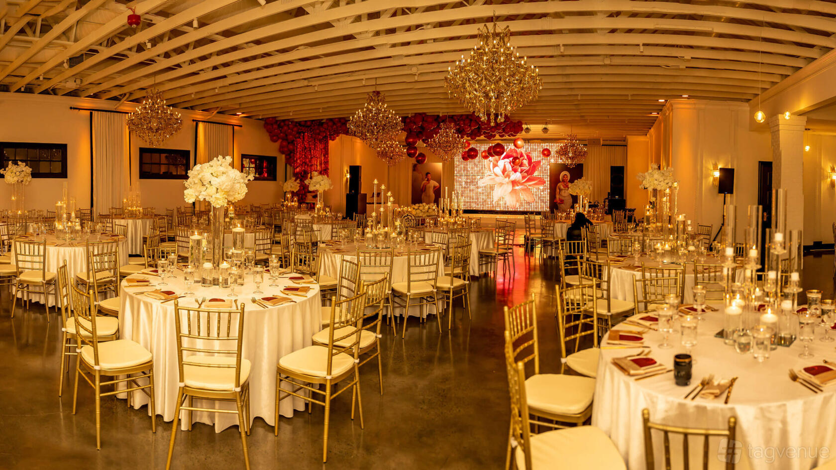 An event venue with round tables draped in white linens, gold chairs, chandeliers, and a large floral wall at Genesis Ballroom.