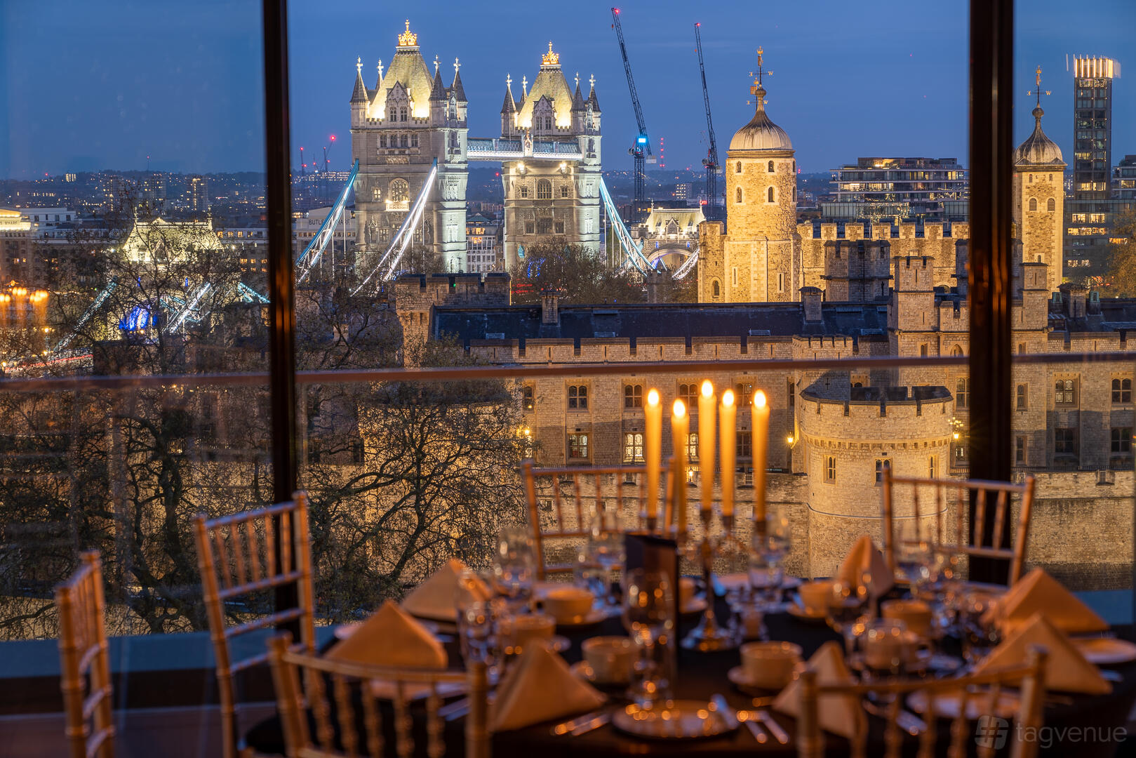 A rooftop venue with floor-to-ceiling windows, round tables set with candles, and Tower Bridge views at Tower Suites by Blue Orchid.