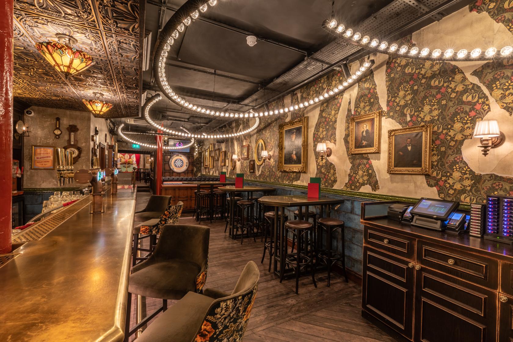 A bar with patterned wallpaper, framed portraits, round tables with stools, and hanging string lights at Flight Club Islington.
