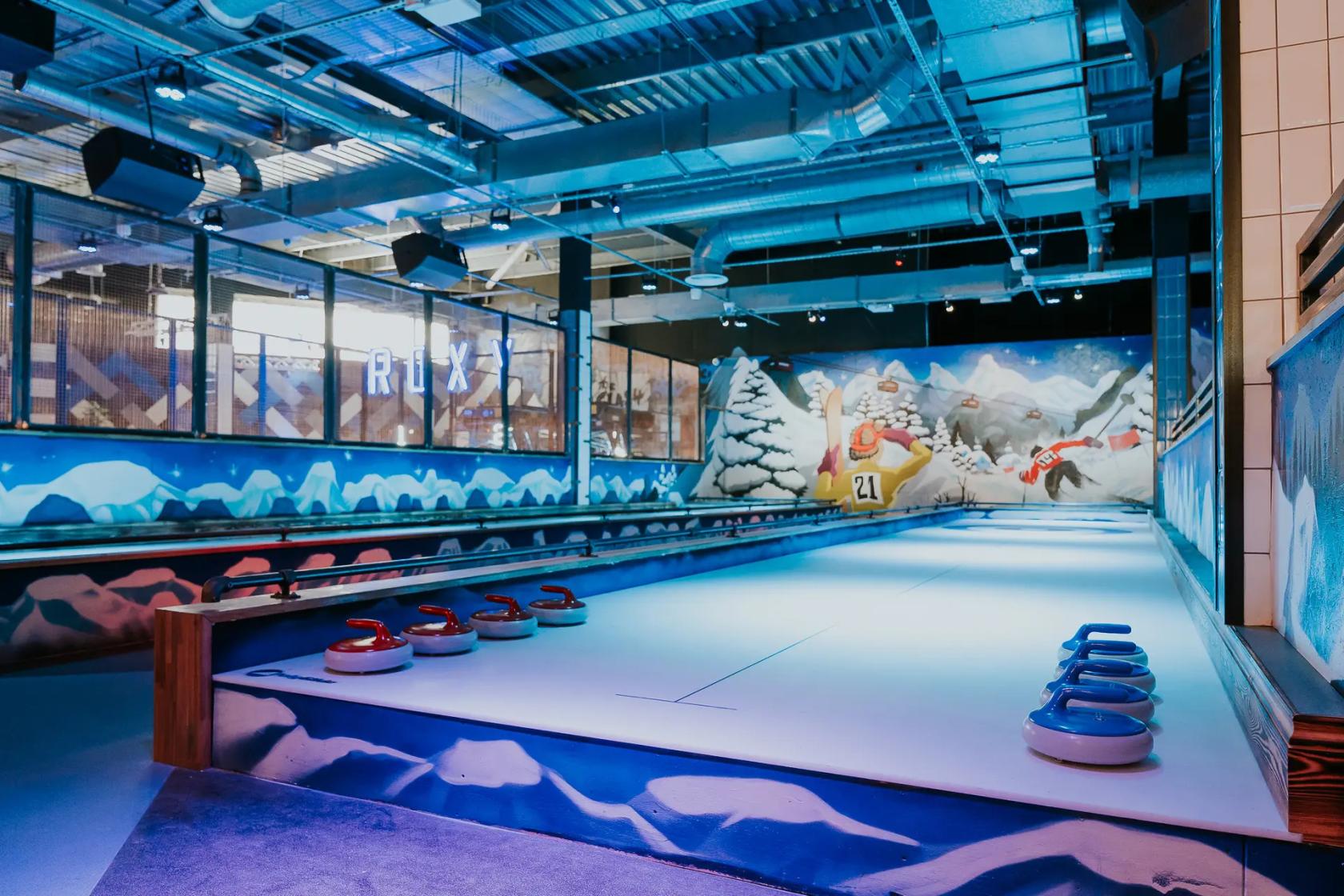A bar with indoor curling lanes, colorful curling stones, and winter-themed mural at Roxy Lanes Bristol Union Street.