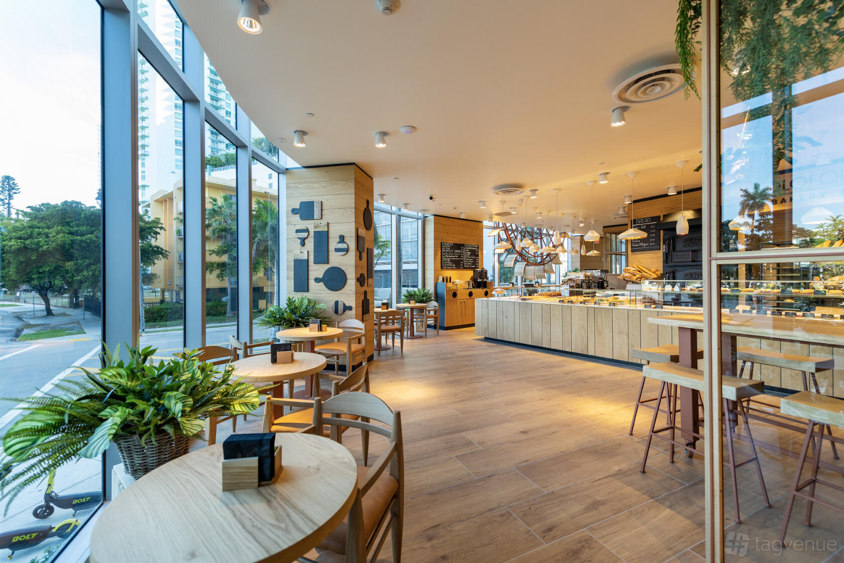 A function room in a cafe with floor-to-ceiling windows, light wooden tables, and indoor plants at Paolo Fontanot Bakery.