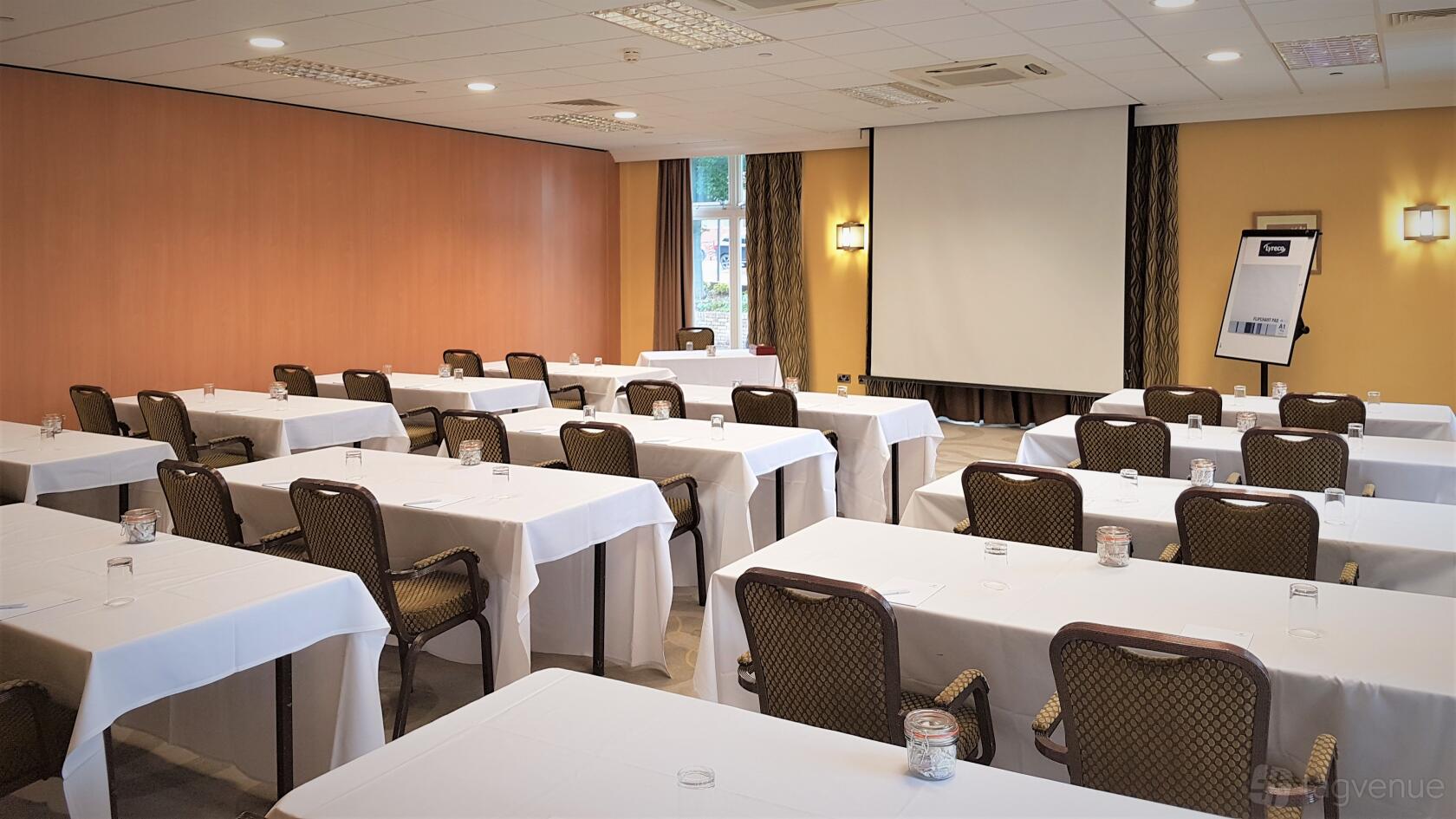 A hotel meeting room with rows of tables draped in white linens, a projector screen, and natural light at Holiday Inn Kenilworth-Warwick.