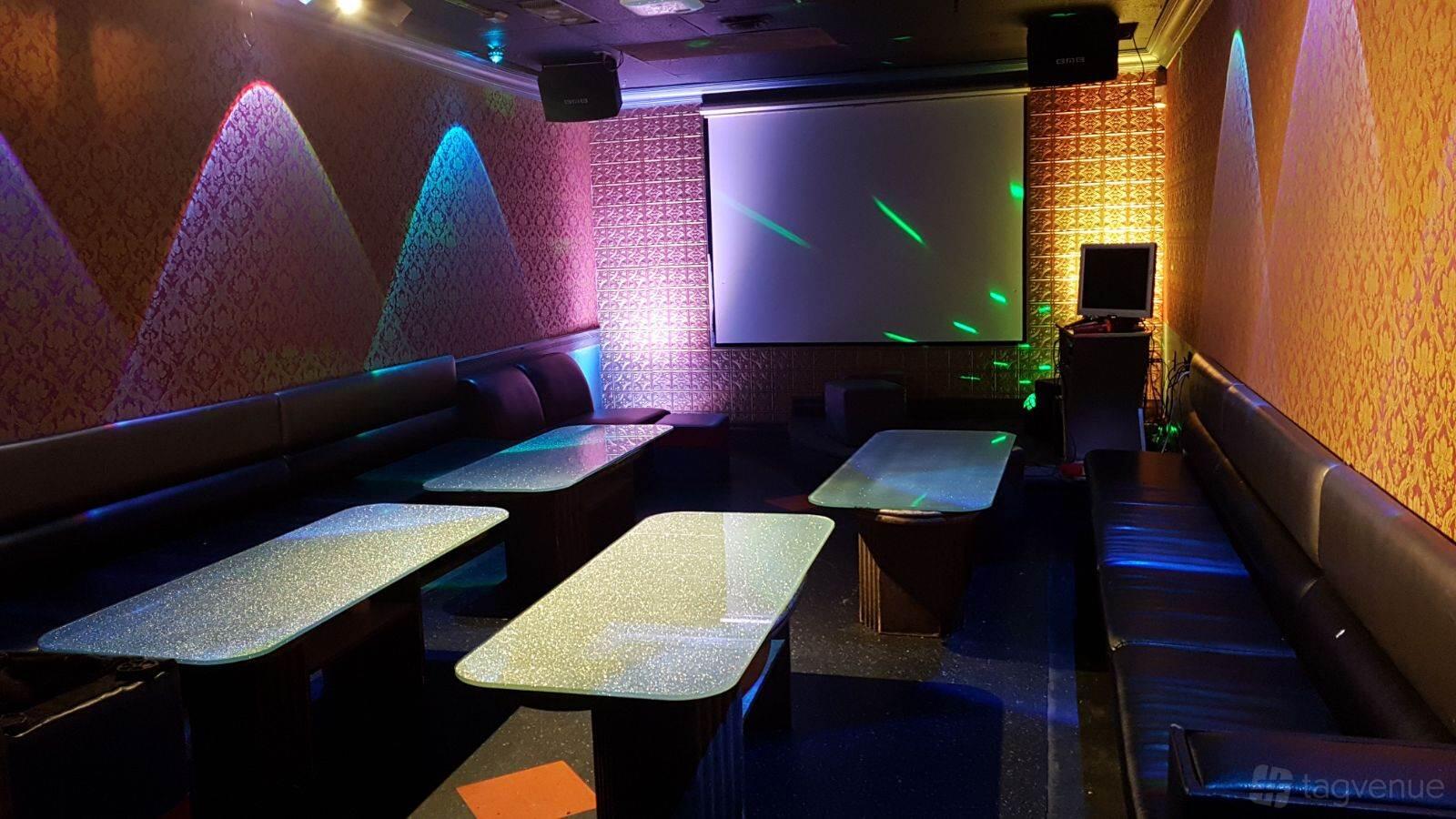 A karaoke bar room with long black sofas, glittery tables, and a large screen at Take One Studios Karaoke.