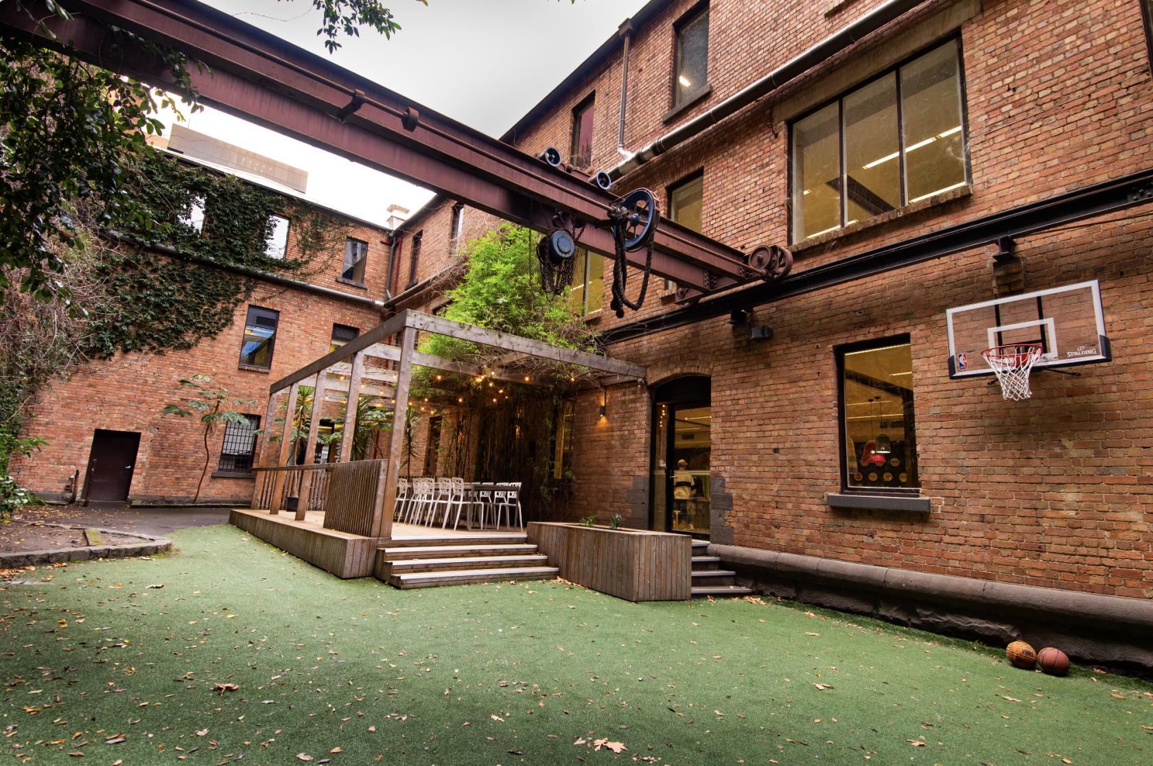 An outdoor courtyard with artificial grass, brick walls, a covered seating area, and a basketball hoop at Stone & Chalk Melbourne.