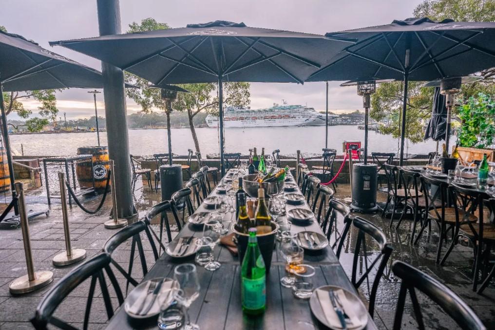A restaurant terrace with long dining tables, black umbrellas, and waterfront views at Elements Bar & Grill Pyrmont.