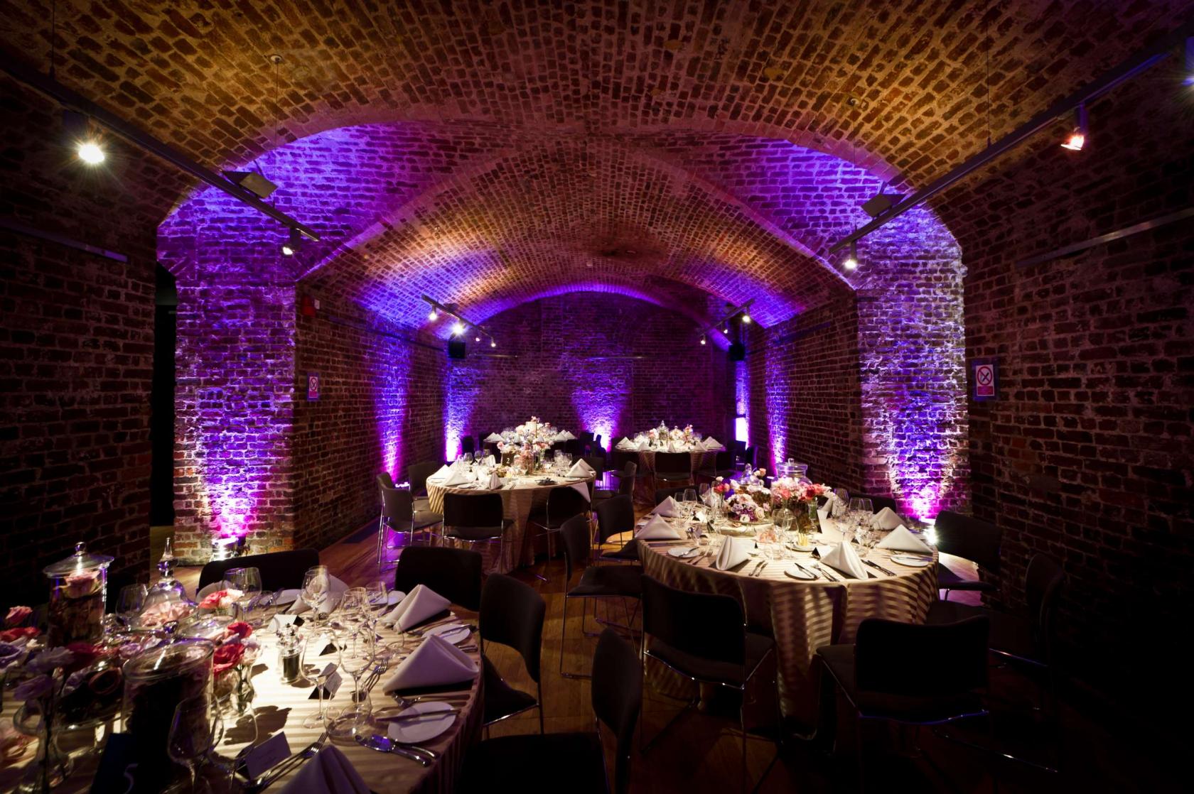 An event space with arched exposed brick ceilings, uplighting, and round tables set for dining at RSA House.
