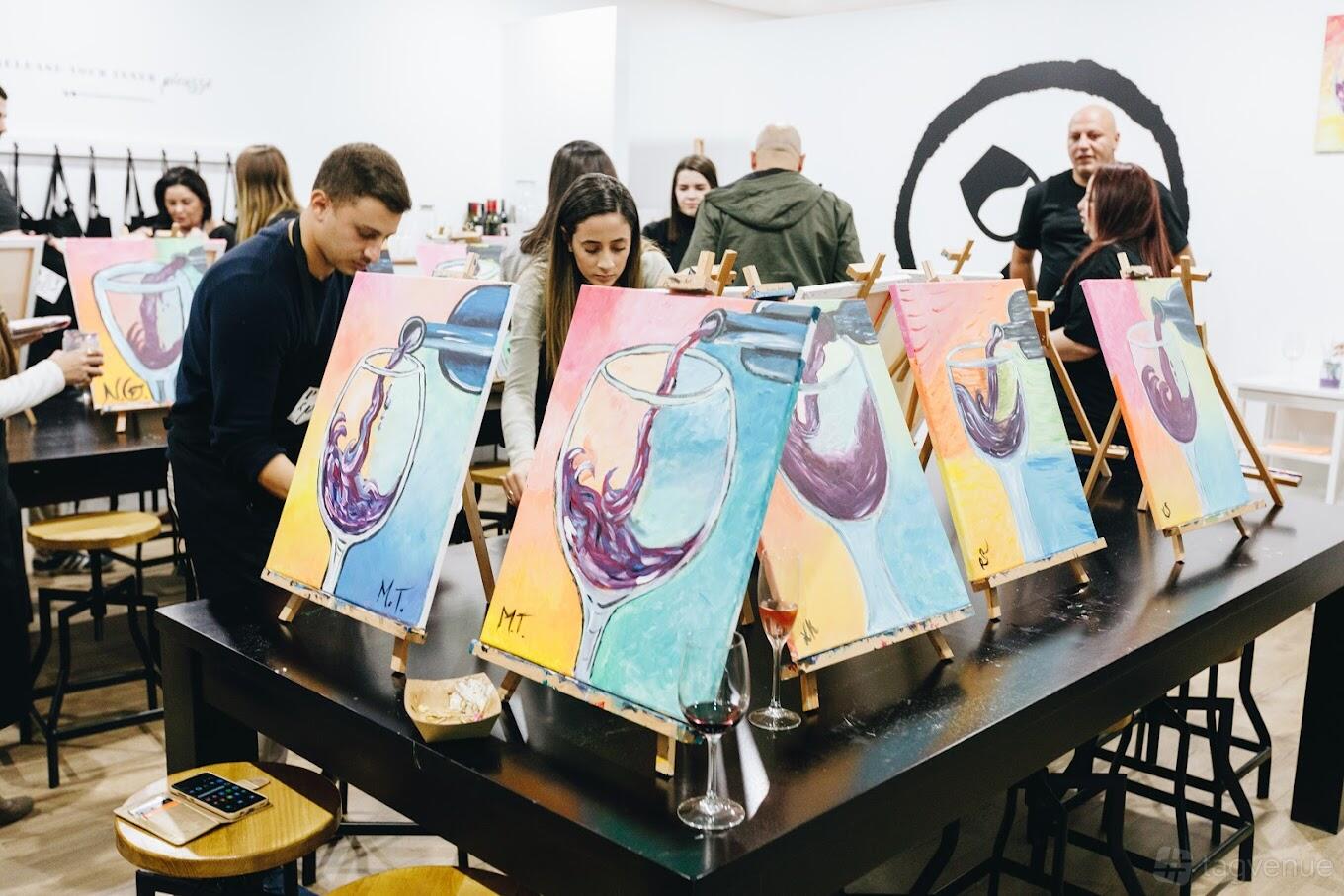 An art studio with long black tables, easels displaying colorful paintings, and people painting at Pinot & Picasso Miranda.