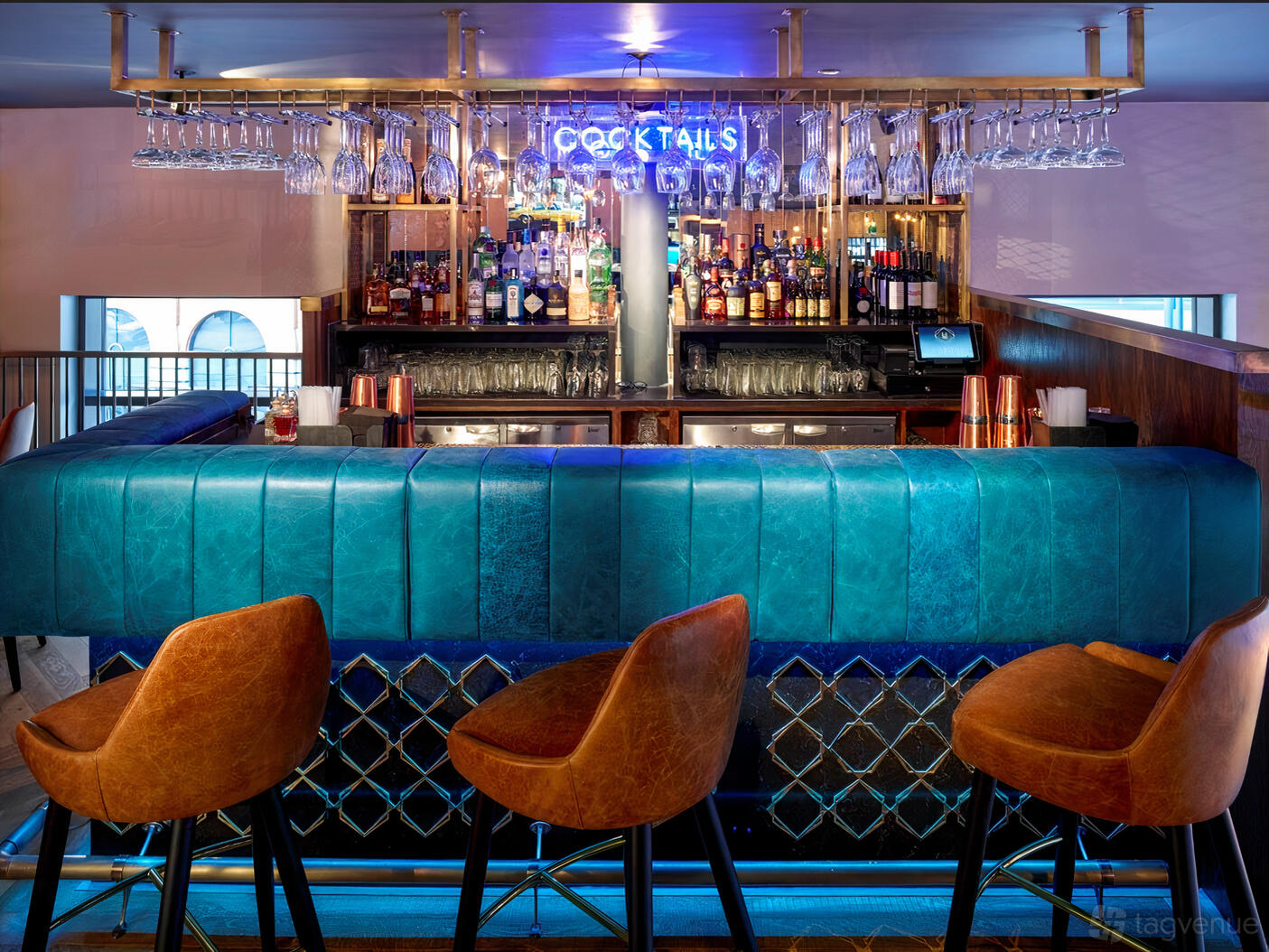 A cocktail bar with a teal tufted bar front, leather stools, hanging glassware, and a neon cocktails sign at Manahatta Greek Street.