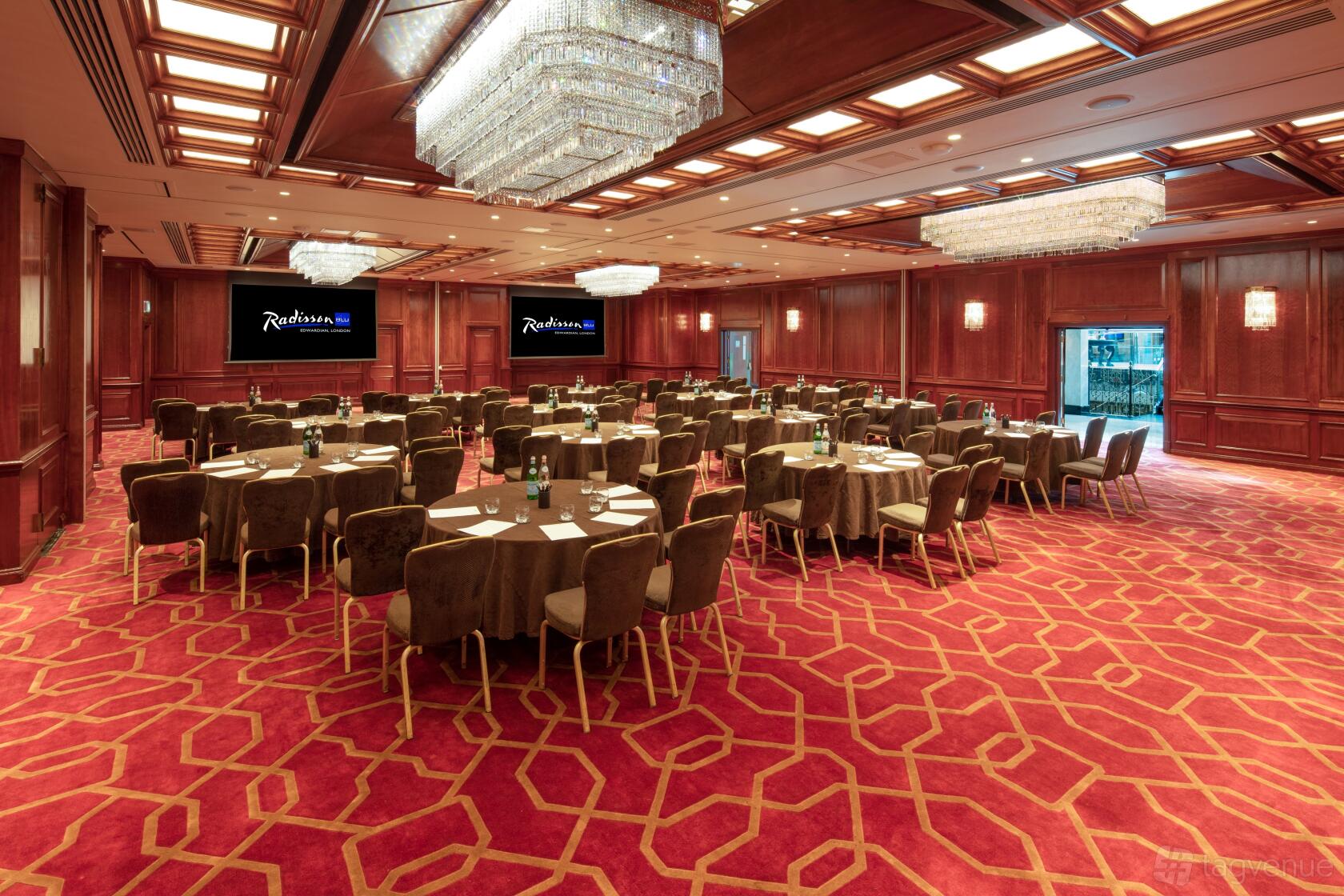 An event space with red patterned carpet, dark chairs, round tables, wood-paneled walls, and chandeliers at Radisson Blu Hotel, London Heathrow.