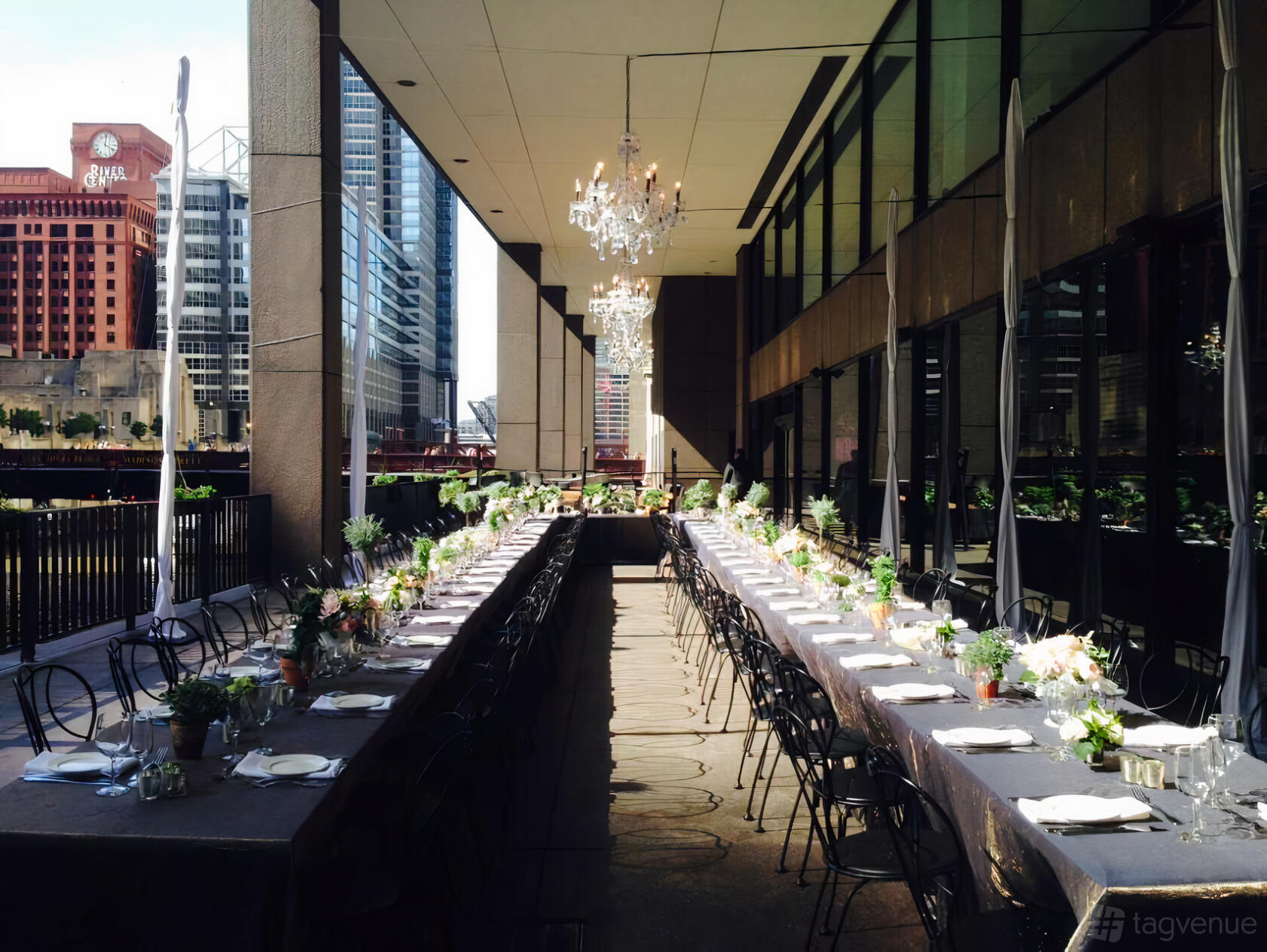 An outdoor restaurant patio with long banquet tables, black chairs, chandeliers, and river views at Rivers Restaurant.