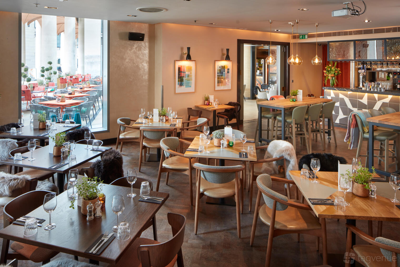 A lounge with wooden tables, cushioned chairs, pendant lights, and a bar area at The Happenstance – Paternoster Square.