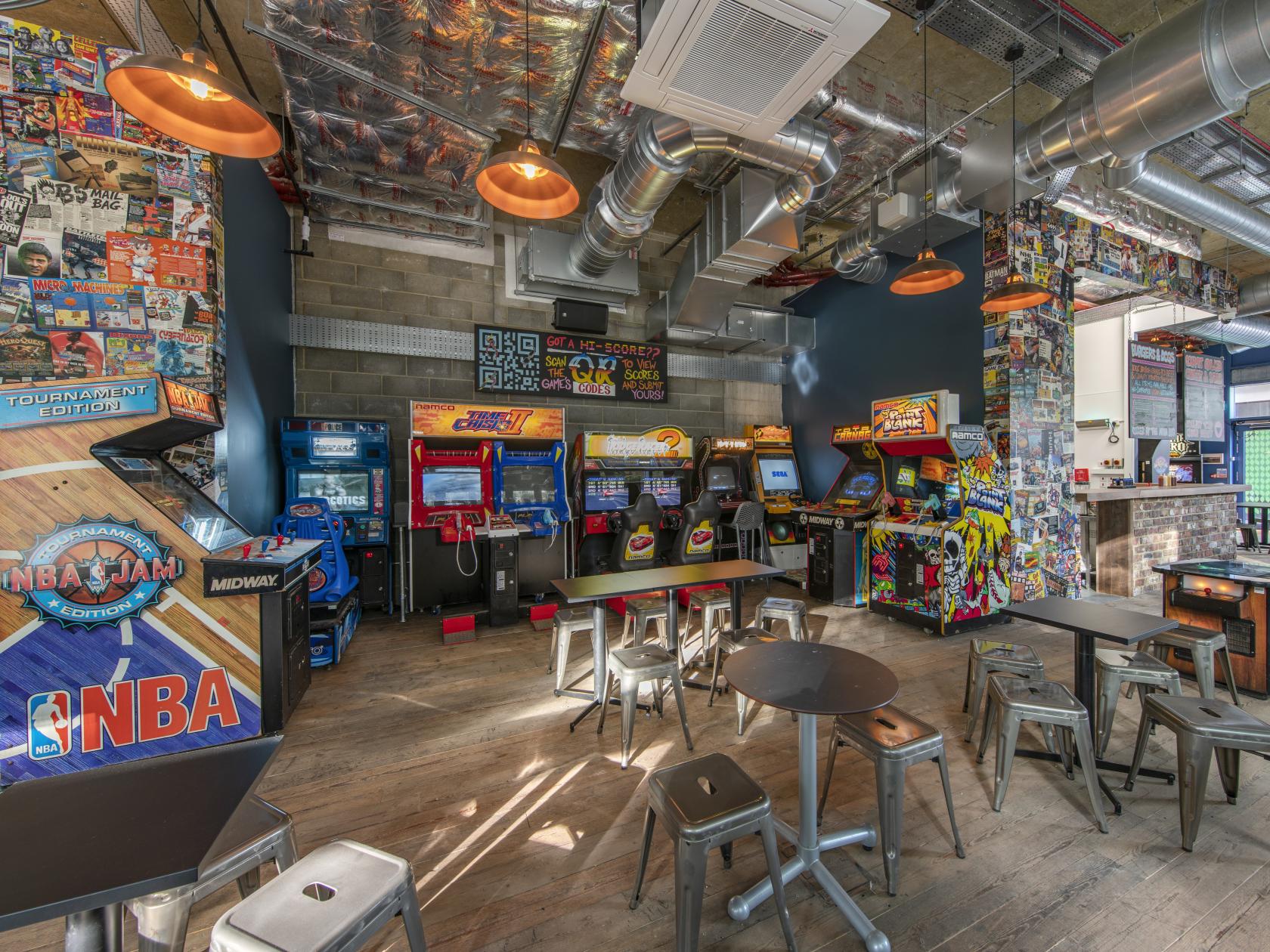 A bar with retro arcade machines, comic-covered walls, and industrial lighting at Four Quarters Elephant Park.