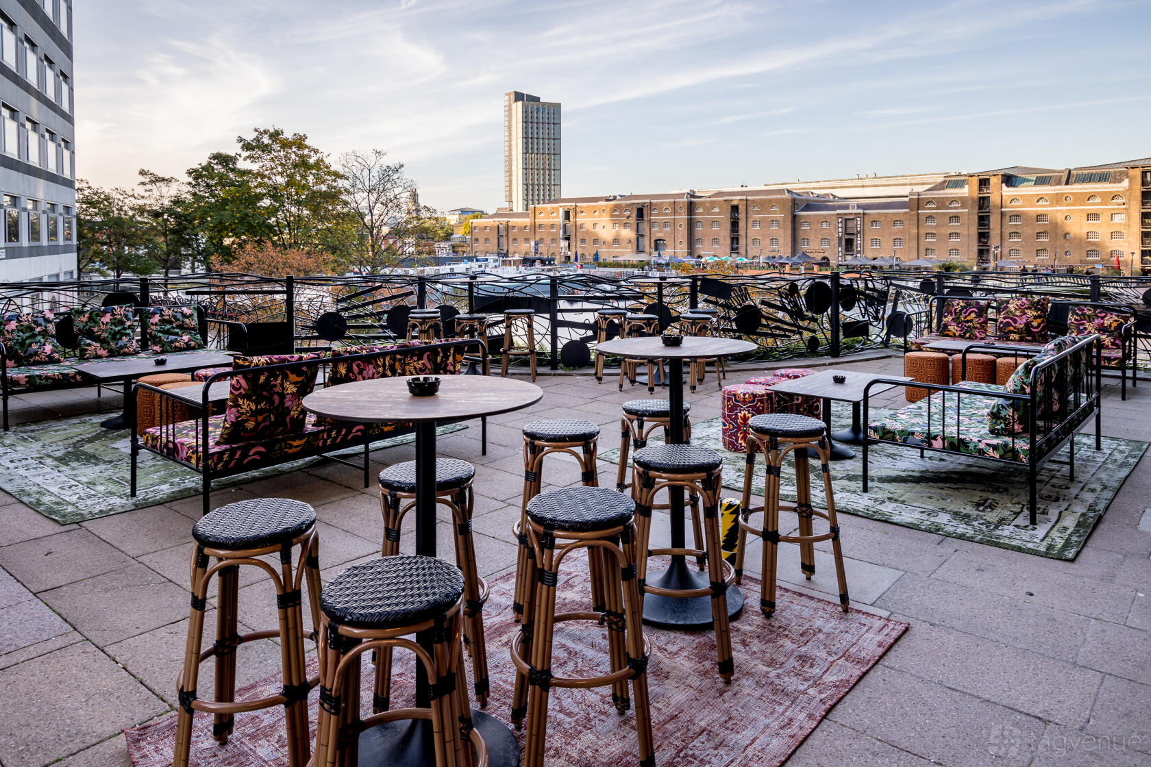 An outdoor cocktail bar terrace with patterned rugs, wicker stools, and city views at The Cocktail Club Canary Wharf.