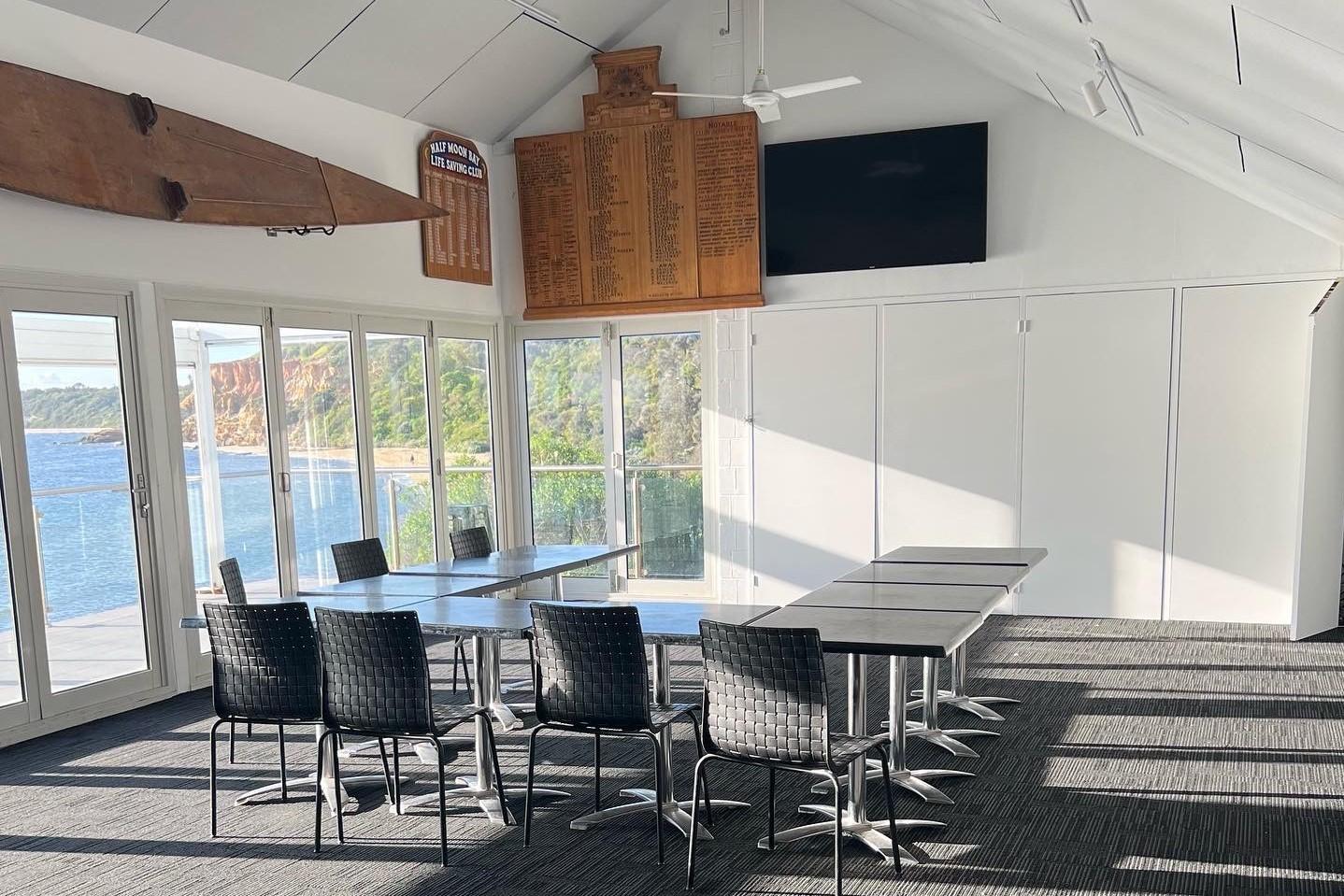 An event venue with floor-to-ceiling windows, ocean views, wall-mounted surfboard, and split tables at Half Moon Bay Surf Life Saving Club.