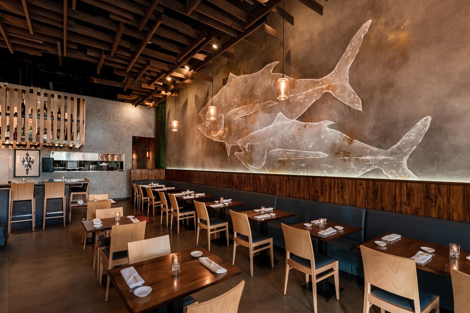 A restaurant dining room with wooden tables, light wood chairs, and a large fish mural on the wall at OMAKAI sushi.