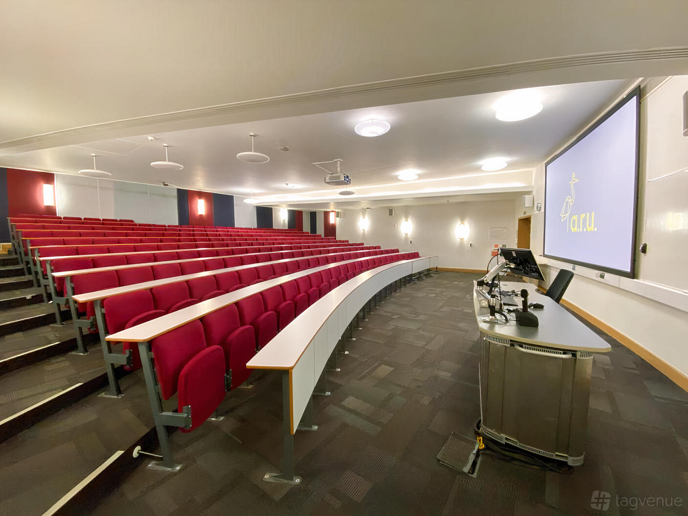 An auditorium with tiered red seating, curved desks, and a projector screen at ARU Venue Hire - Cambridge.
