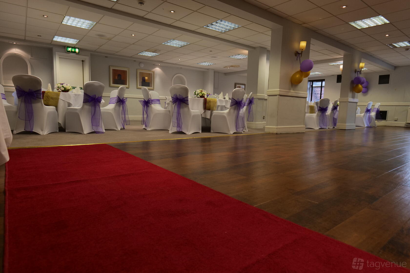 A banquet hall with wood flooring, red carpet, and white chairs decorated with purple sashes at The Great Barr Hotel.
