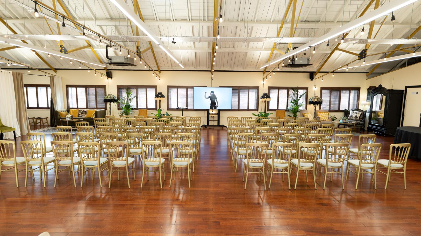 A spacious event hall with wooden floors, rows of gold chairs, large windows with shutters, and a projector screen at Lumiere London Old Street.