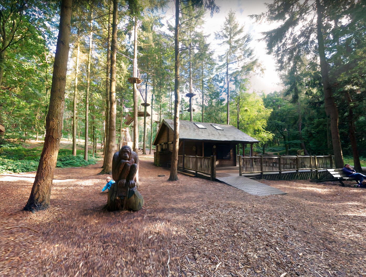 An outdoor venue with tall pine trees, a wooden cabin with a deck, and a carved monkey sculpture at Go Ape Wyre.