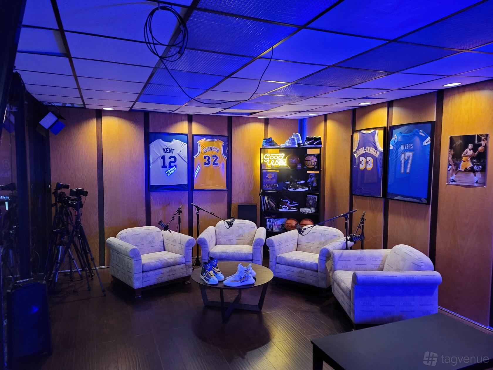 A podcast studio with four armchairs, microphones, sports jerseys on wood-paneled walls, and blue ambient lighting at Infanity Studios.