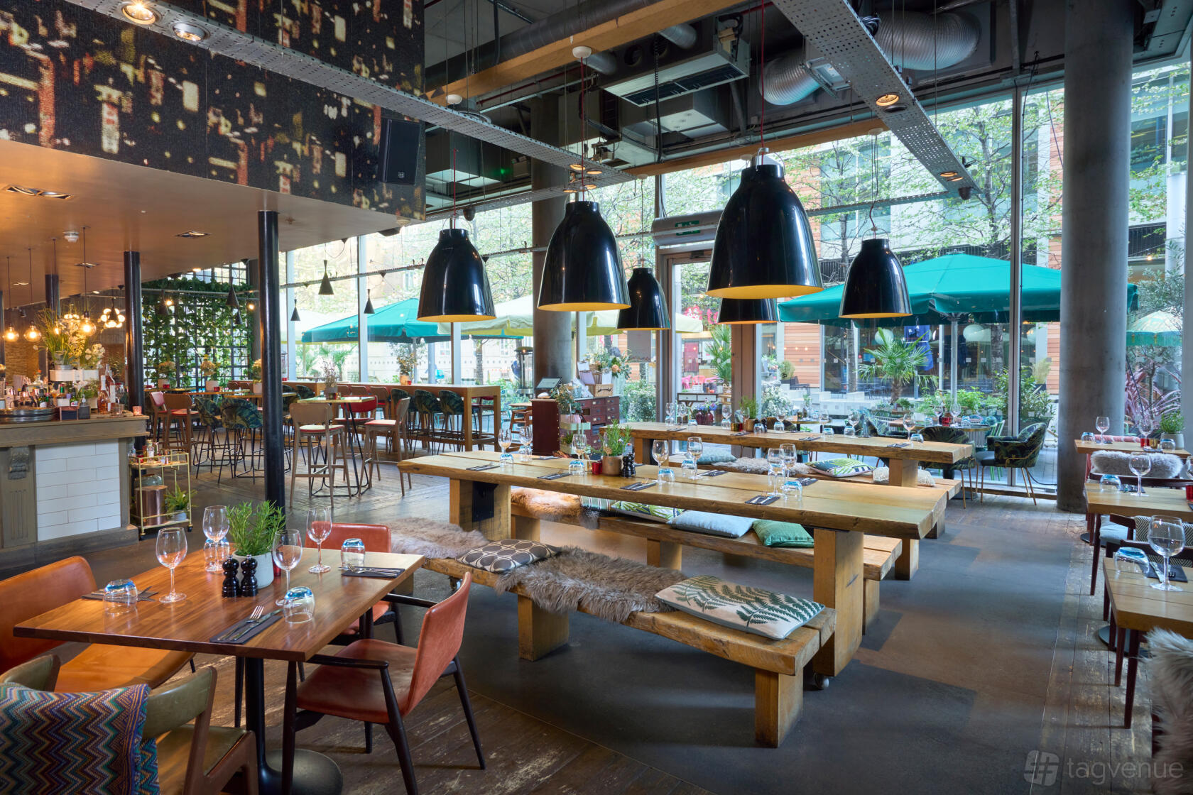 A restaurant with floor-to-ceiling windows, long wooden tables, pendant lighting, and fur bench seating at The Refinery Bankside.