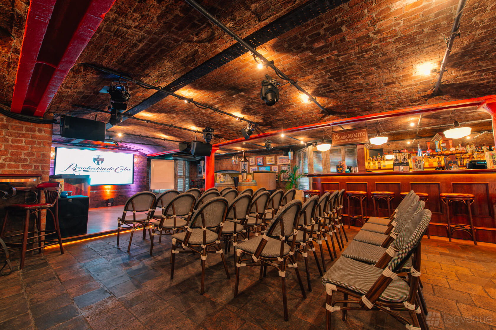 An event space in a bar with exposed brick ceilings, rows of chairs, a stage, and a lit bar at Revolucion de Cuba Liverpool.
