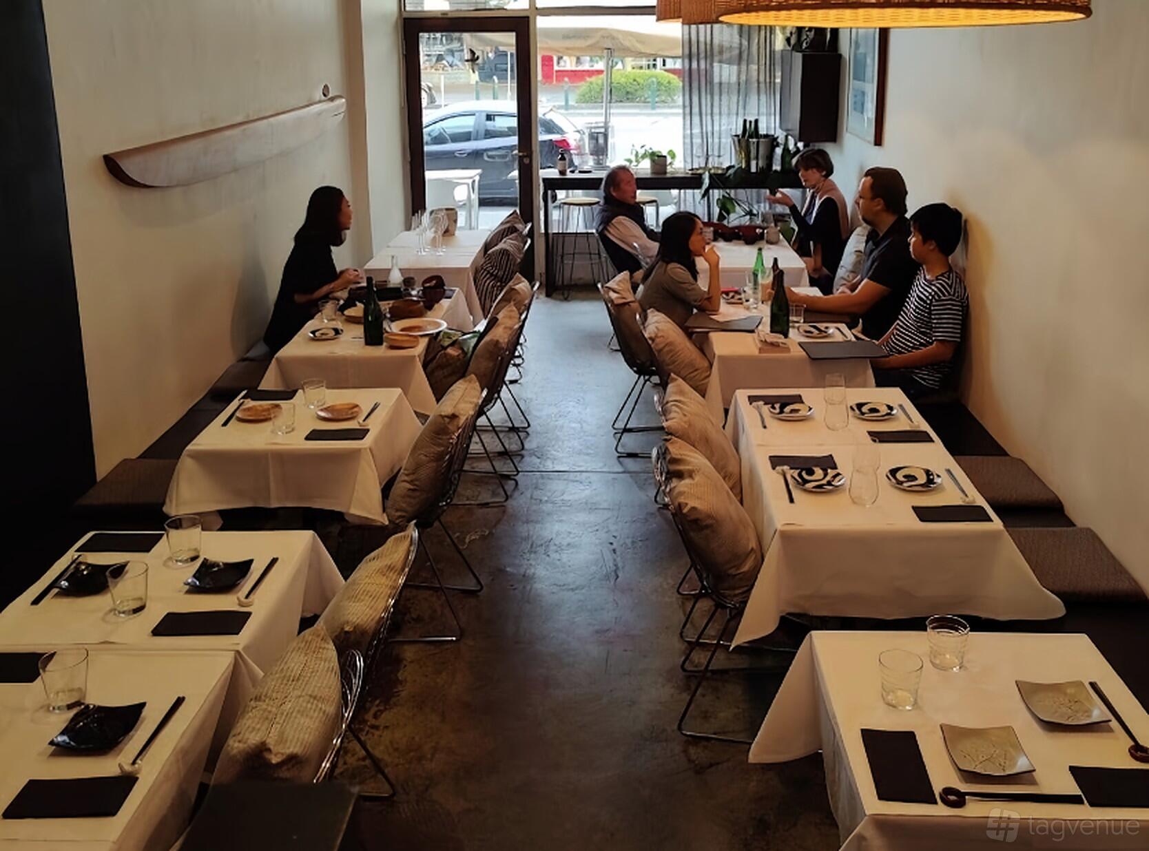 A restaurant dining area with tables set with black and white dinnerware, cushioned chairs, and large windows at Eis Japanese Restaurant.