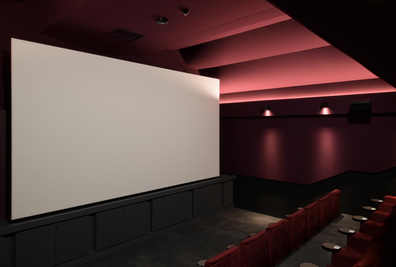 A cinema screening room with red tiered seating, a large white screen, and wall sconces at Curzon Camden.