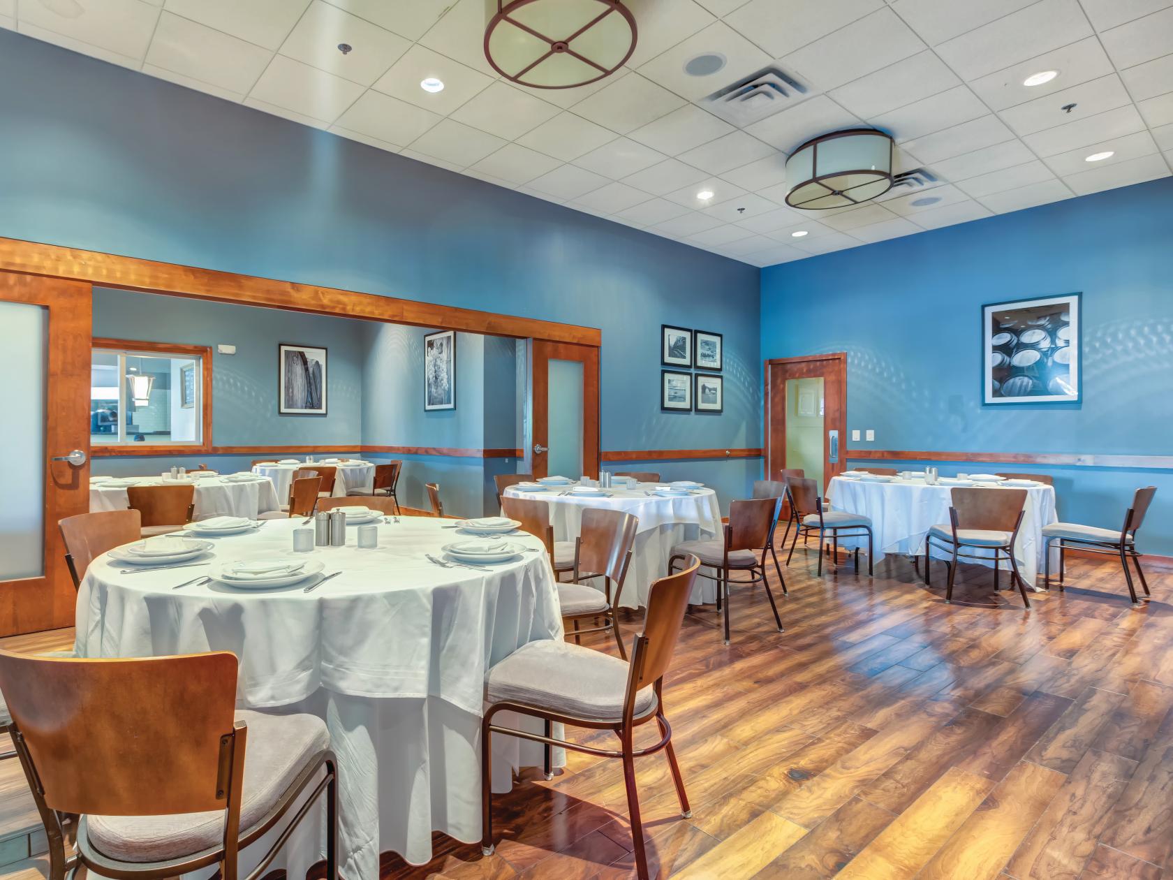 A dining room with round tables draped in white linens, wooden chairs, framed art, and hardwood floors at Pinstripes - Oakbrook.