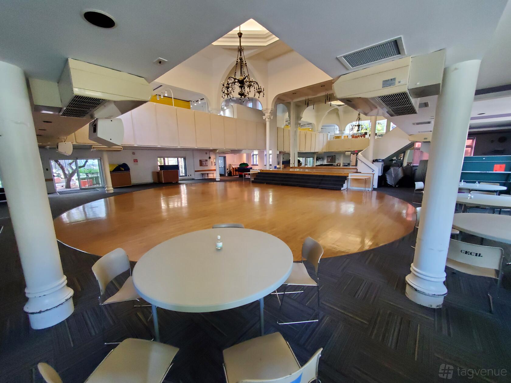 An auditorium with a polished wood stage, high ceilings, chandeliers, and round tables at Cecil Community Centre.