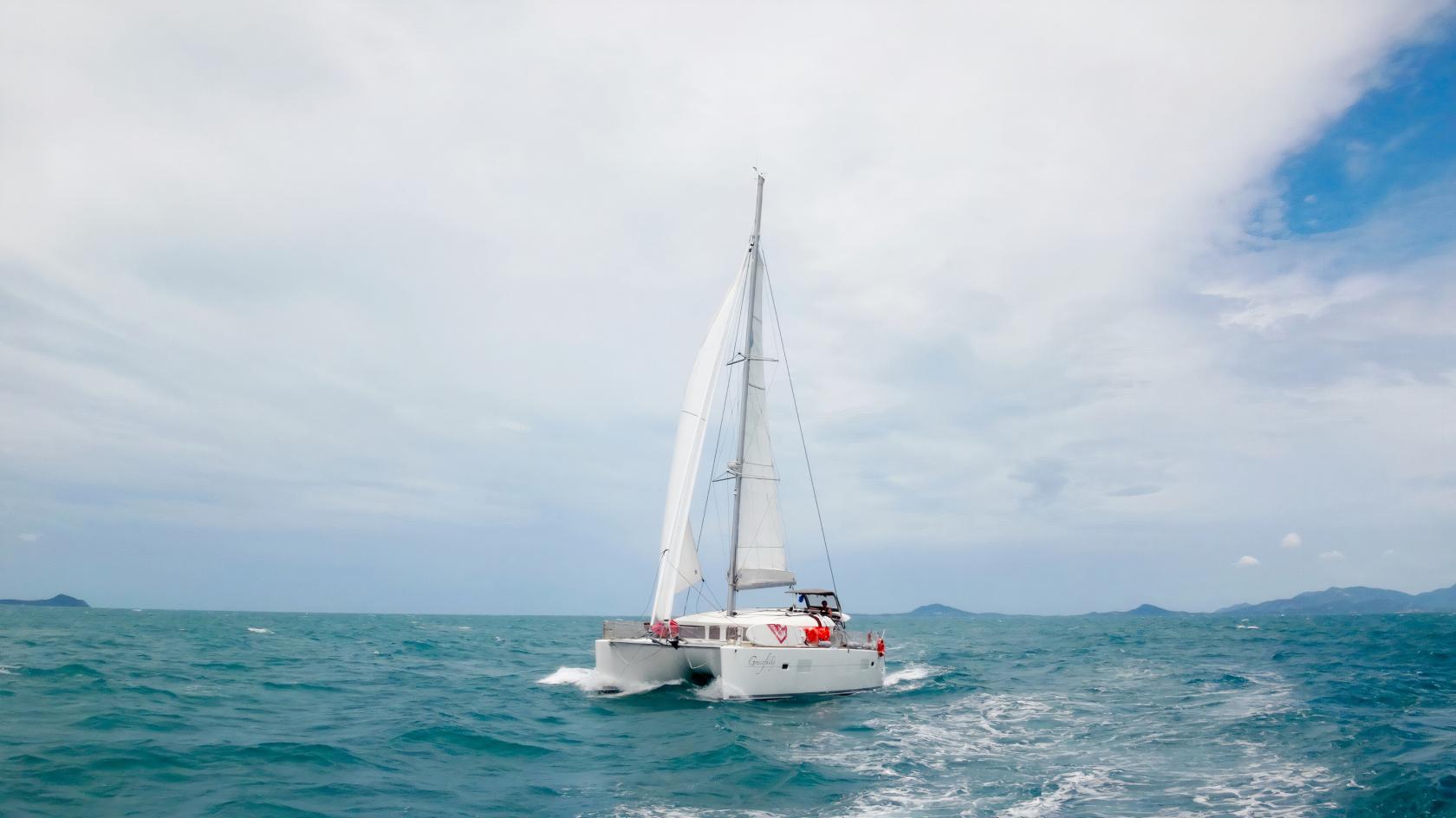 A sailing boat with white sails cruising on open blue water near distant islands at Ximula Sail.