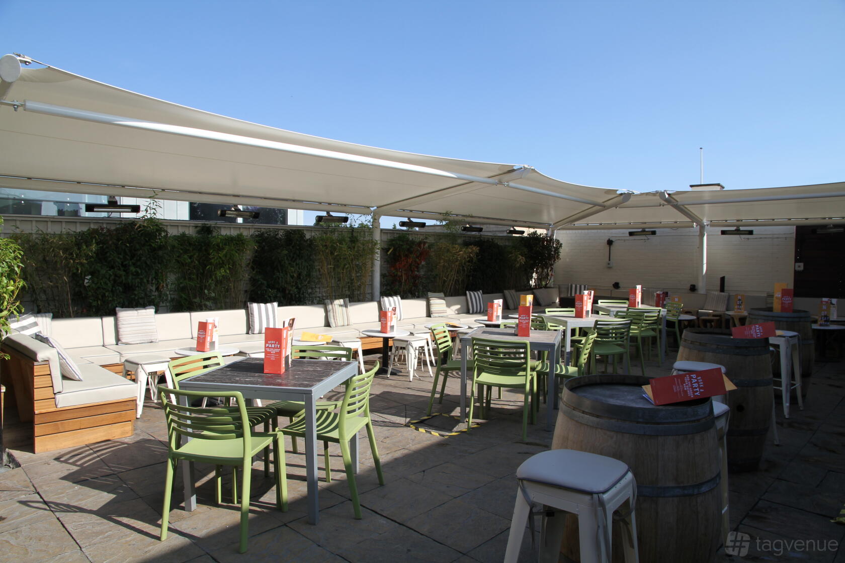 A rooftop terrace with shade canopies, green chairs, long benches, and potted greenery at Revolution Cambridge.