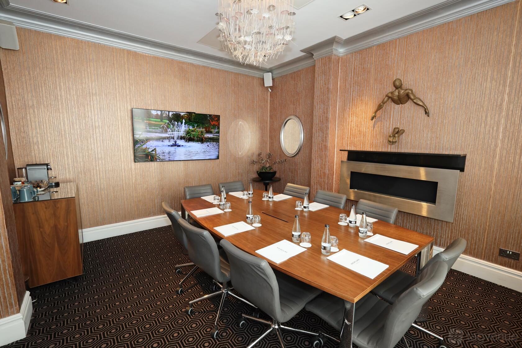 An event space in a hotel with a wood conference table, leather chairs, chandelier, and wall-mounted TV at Mercure London Hyde Park.