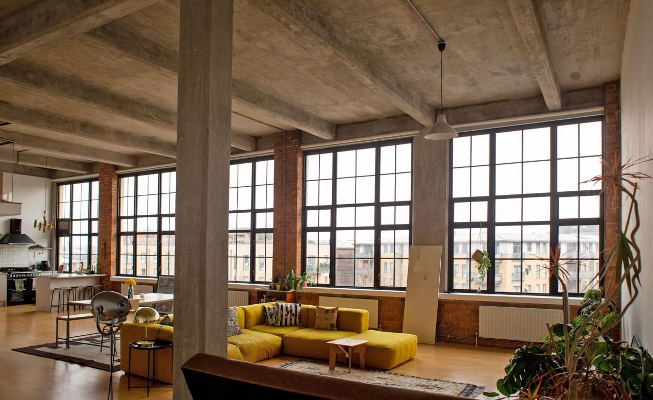 A photo studio with floor-to-ceiling industrial windows, exposed concrete ceiling, and a yellow sectional sofa at Dehav Studios.