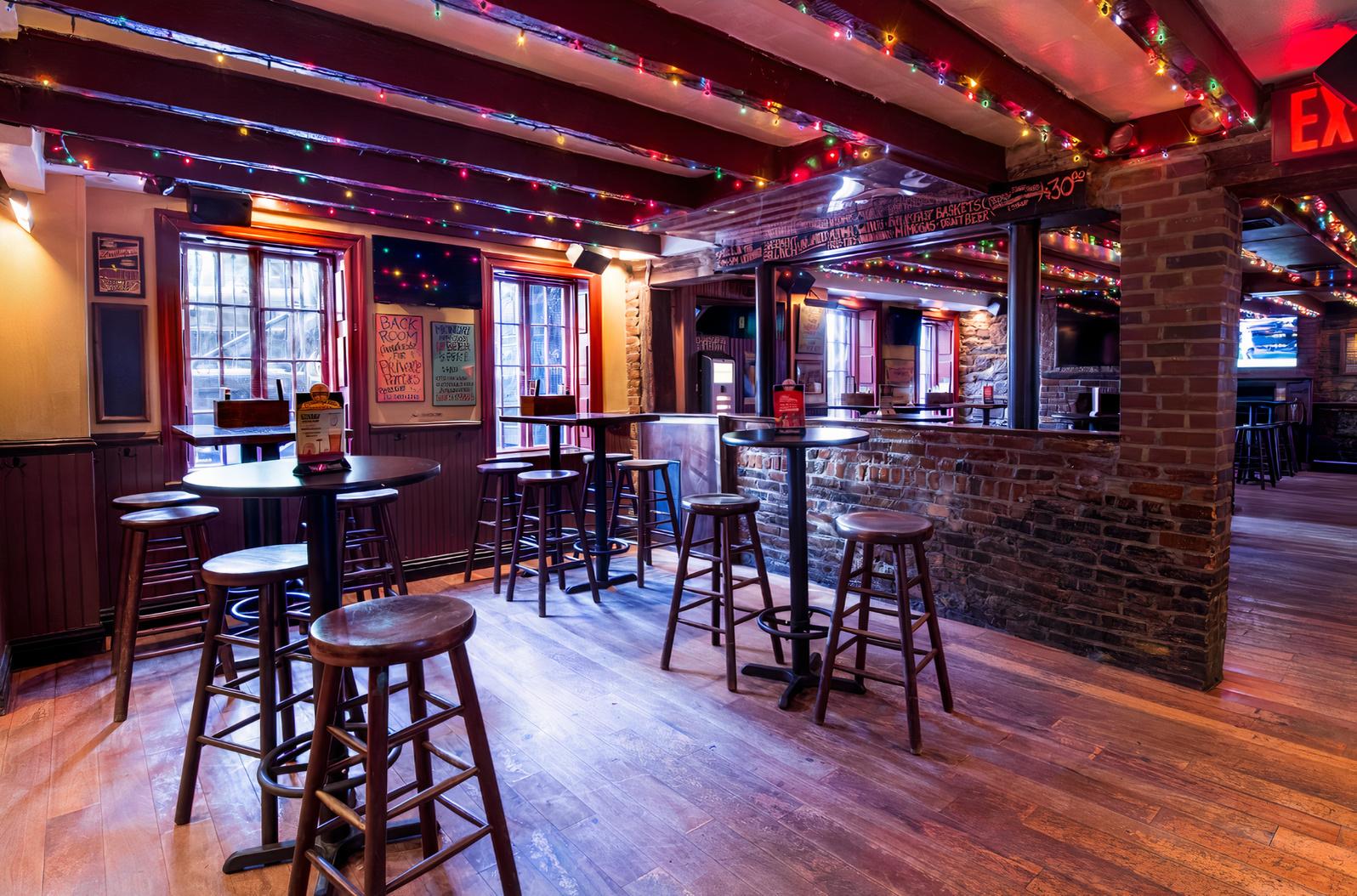 A bar with exposed brick, string lights along wood beam ceilings, and tall tables with stools at Down the Hatch.