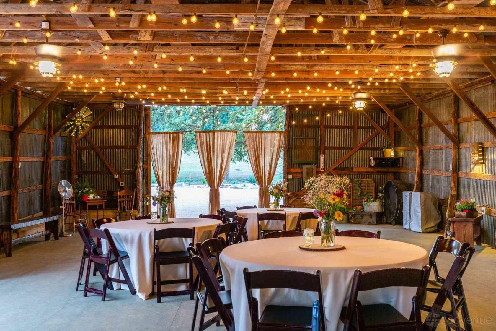 A rustic barn with exposed wooden beams, string lights, round tables with linens, and open barn doors at Texas Rock House.